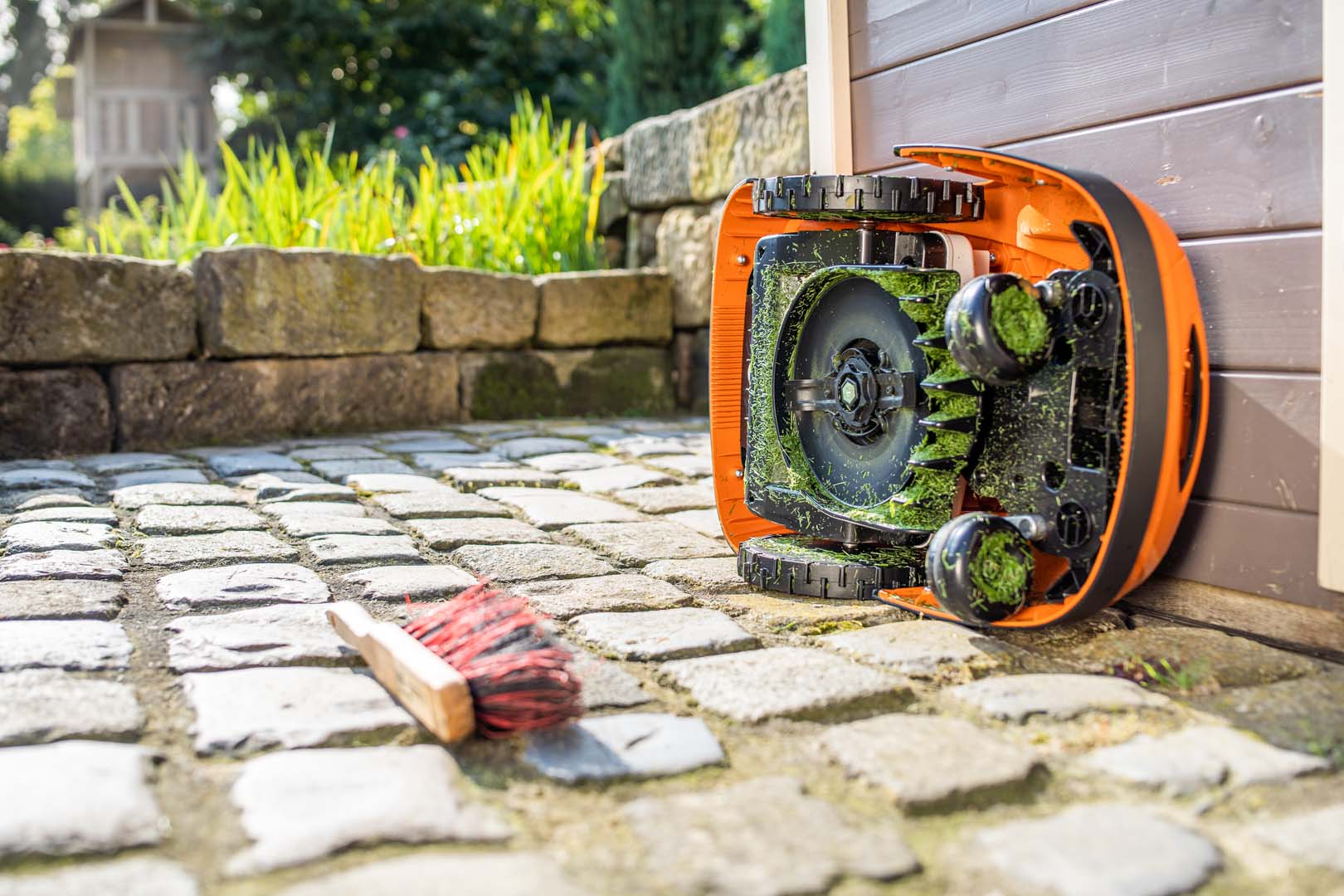 A STIHL iMOW® robot lawn mower turned on its side ready for cleaning, with a brush on the ground nearby